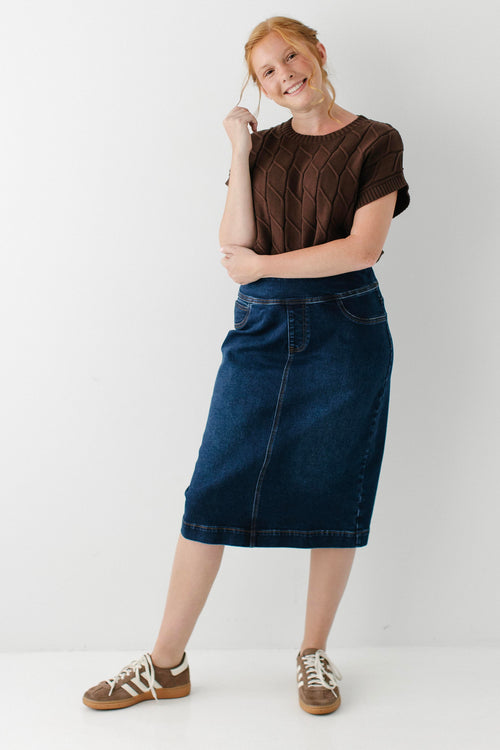 A woman modeling a dark brown short-sleeve knit top and a dark blue denim midi skirt, paired with brown and white sneakers, against a plain white studio background.