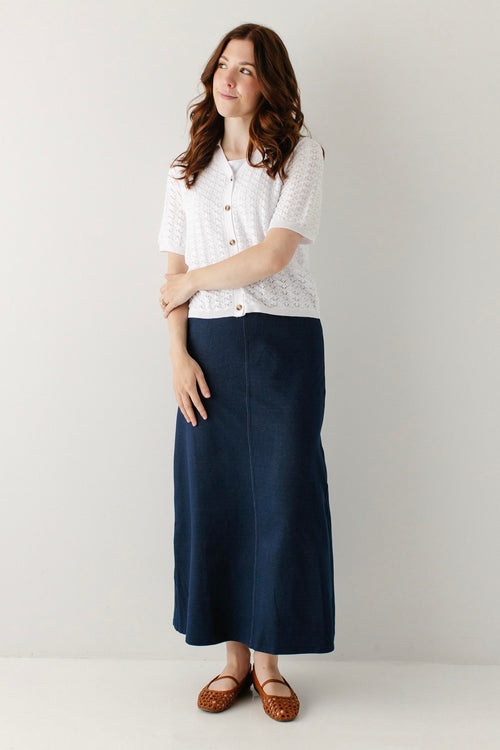 A woman modeling a white short-sleeve knit top with buttons and a dark navy blue midi skirt, paired with brown flat shoes, against a light gray studio background.